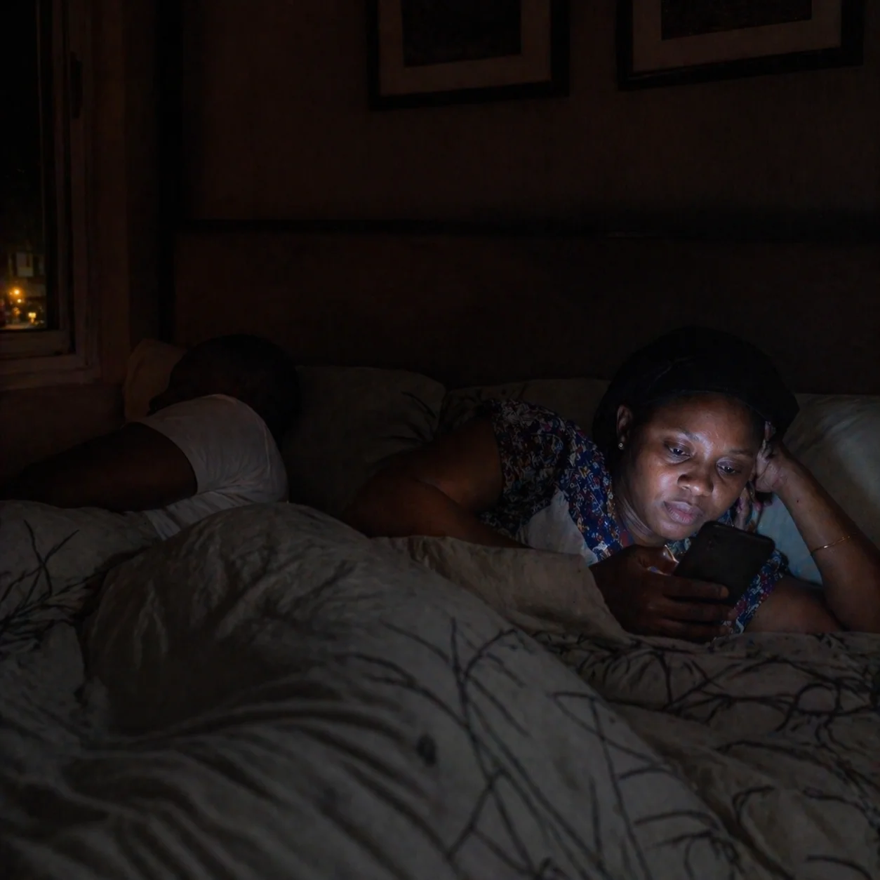 Adaeze in bed reading her phone while David sleeps, expression carefully neutral