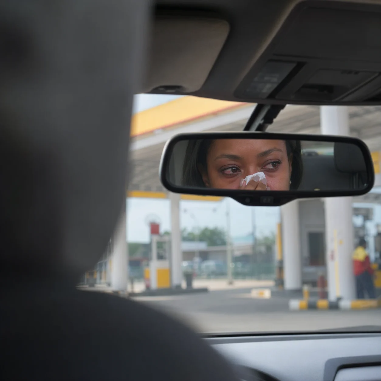 Adaeze's reflection in the rear-view mirror at a filling station, pressing a tissue to the bridge of her nose