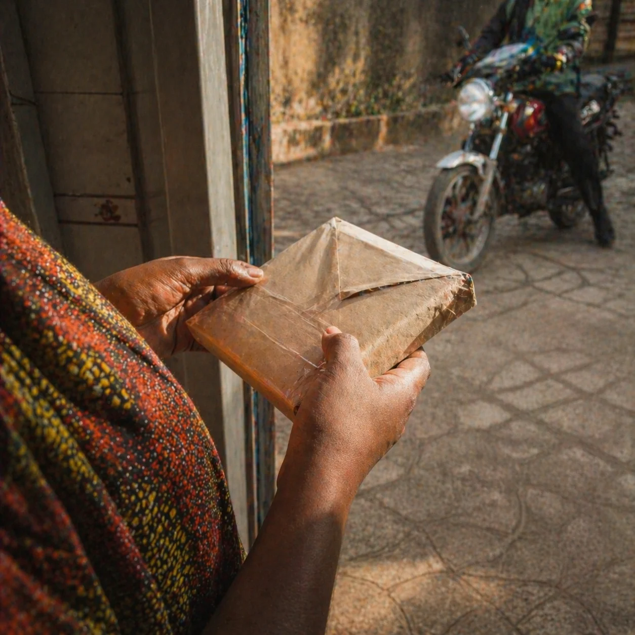 Adaeze's hands turning a plain brown parcel at the doorstep, delivery rider behind