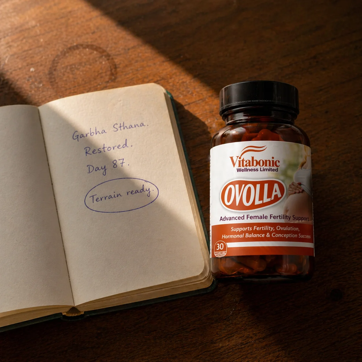 Ovolla bottle on worn Lagos desk, notebook beside it with Garbha Sthana Restored Day 87